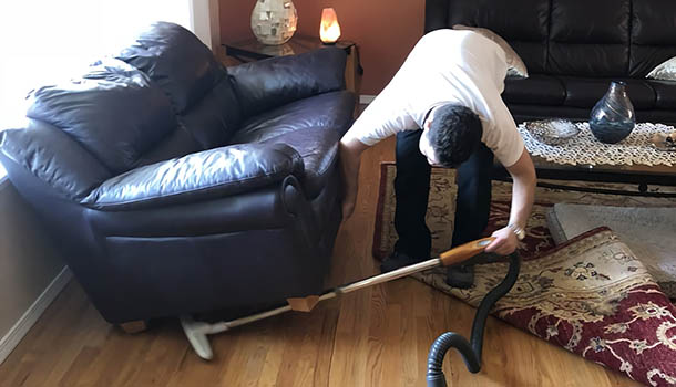 A man vacuuming a couch in a tidy living room, ensuring cleanliness and comfort in his home environment.